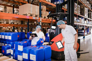 Warehouse team managing inventory and logistics Two employees use tablets while inspecting blue containers in a warehouse. Shelving units filled with pallets and packaged goods are visible throughout the storage area.