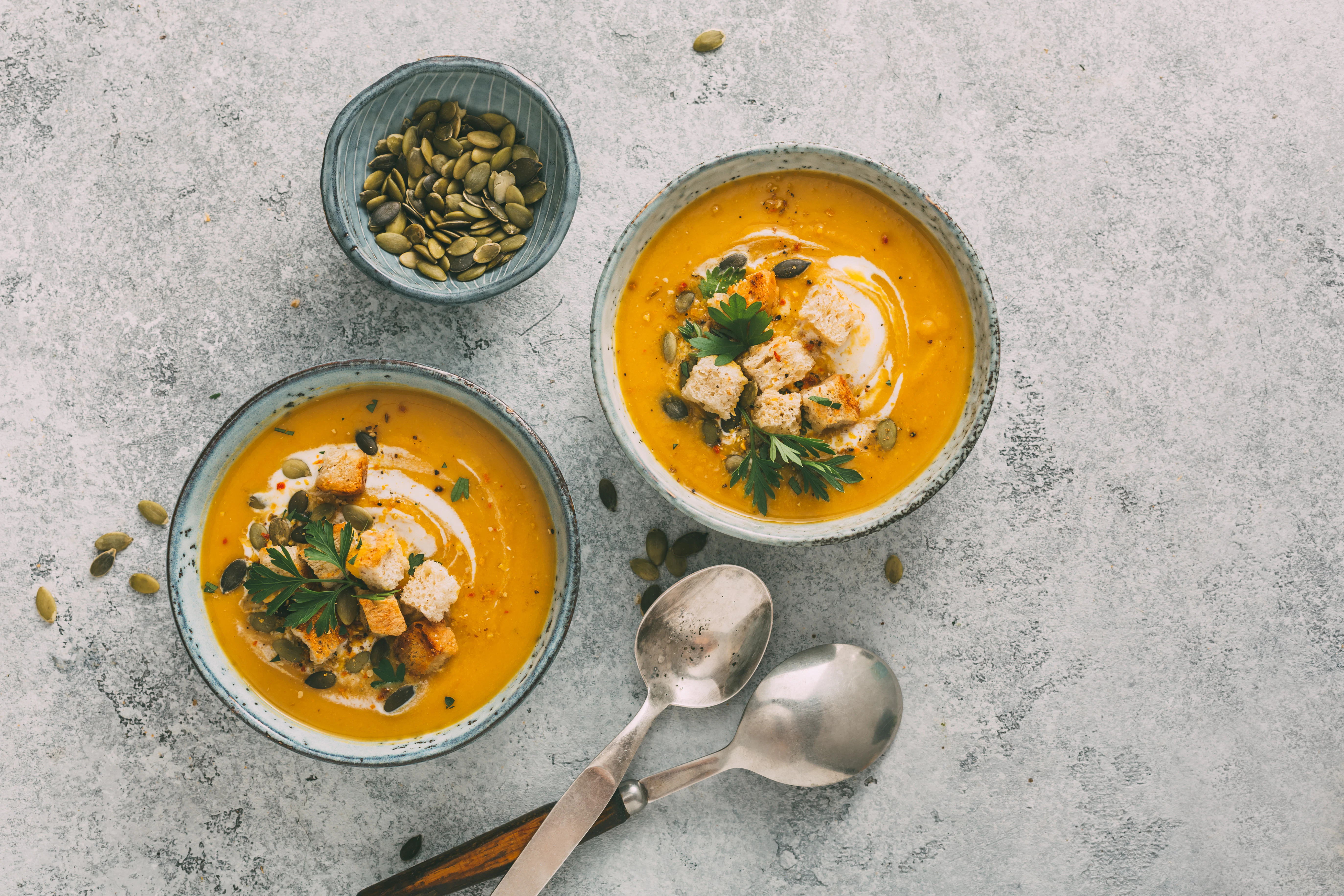 Creamy pumpkin soup with herbs and seeds Two bowls of orange pumpkin soup are placed on a light surface and garnished with cream, croutons, parsley, and pumpkin seeds. A small bowl of seeds and two spoons are arranged nearby.