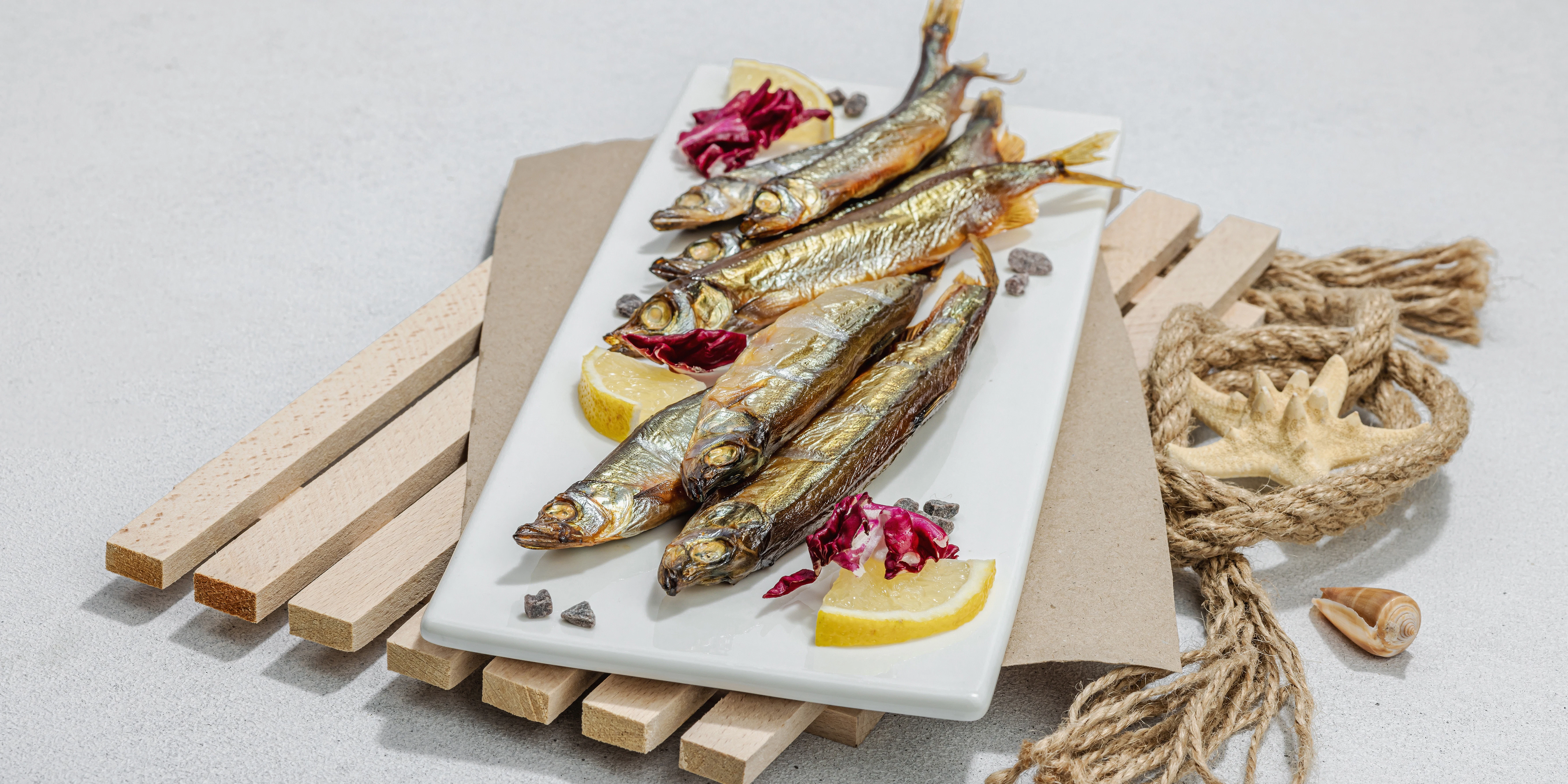 Small fish served with lemon and herbs Several whole small fish are arranged on a rectangular white plate with lemon wedges and red cabbage leaves. The plate is placed on wooden boards with rope decoration in the background.