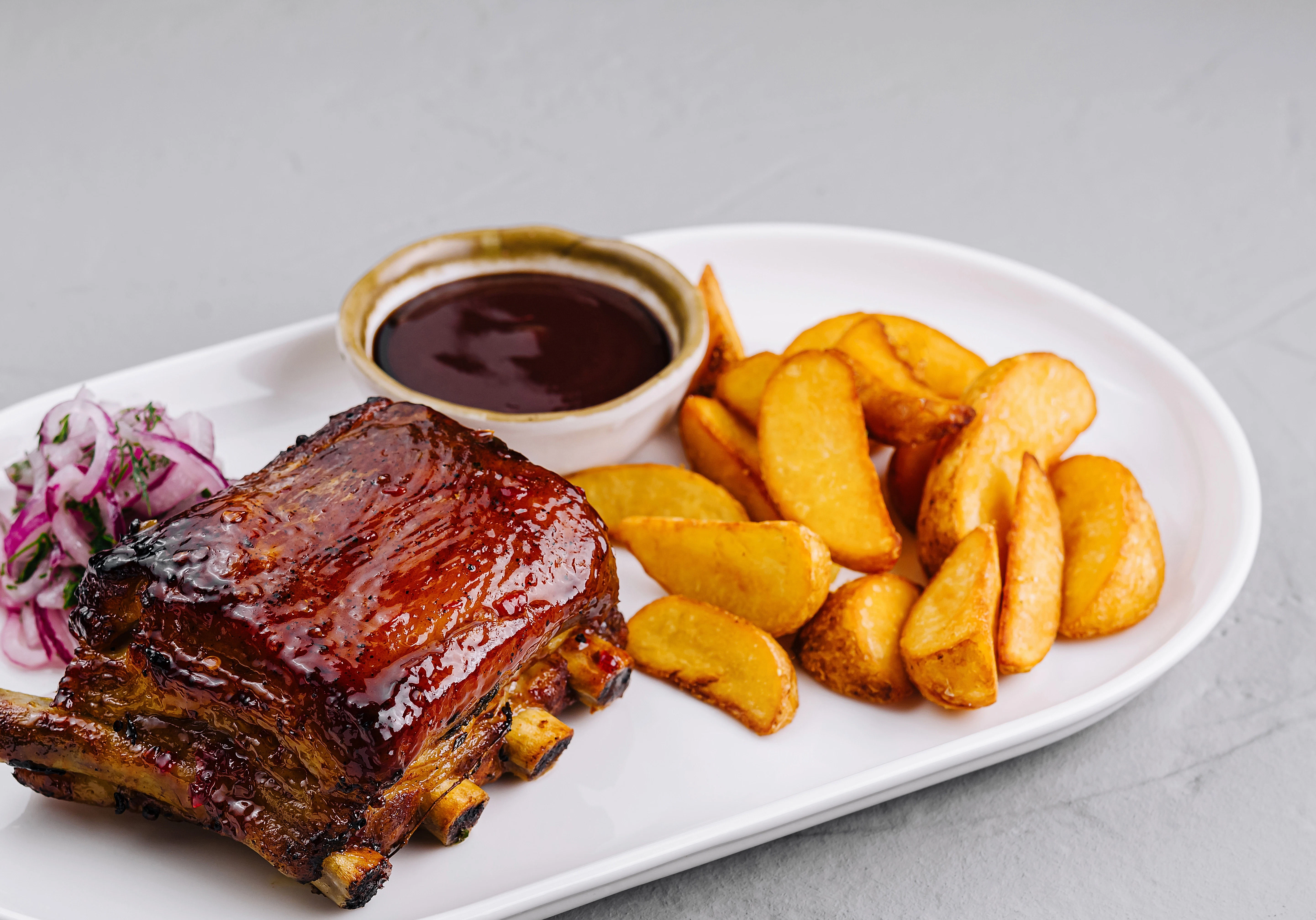Glazed ribs with roasted potato wedges A portion of glazed ribs is served on a white plate with golden potato wedges and a small bowl of dark sauce. Pickled onions are placed next to the meat.