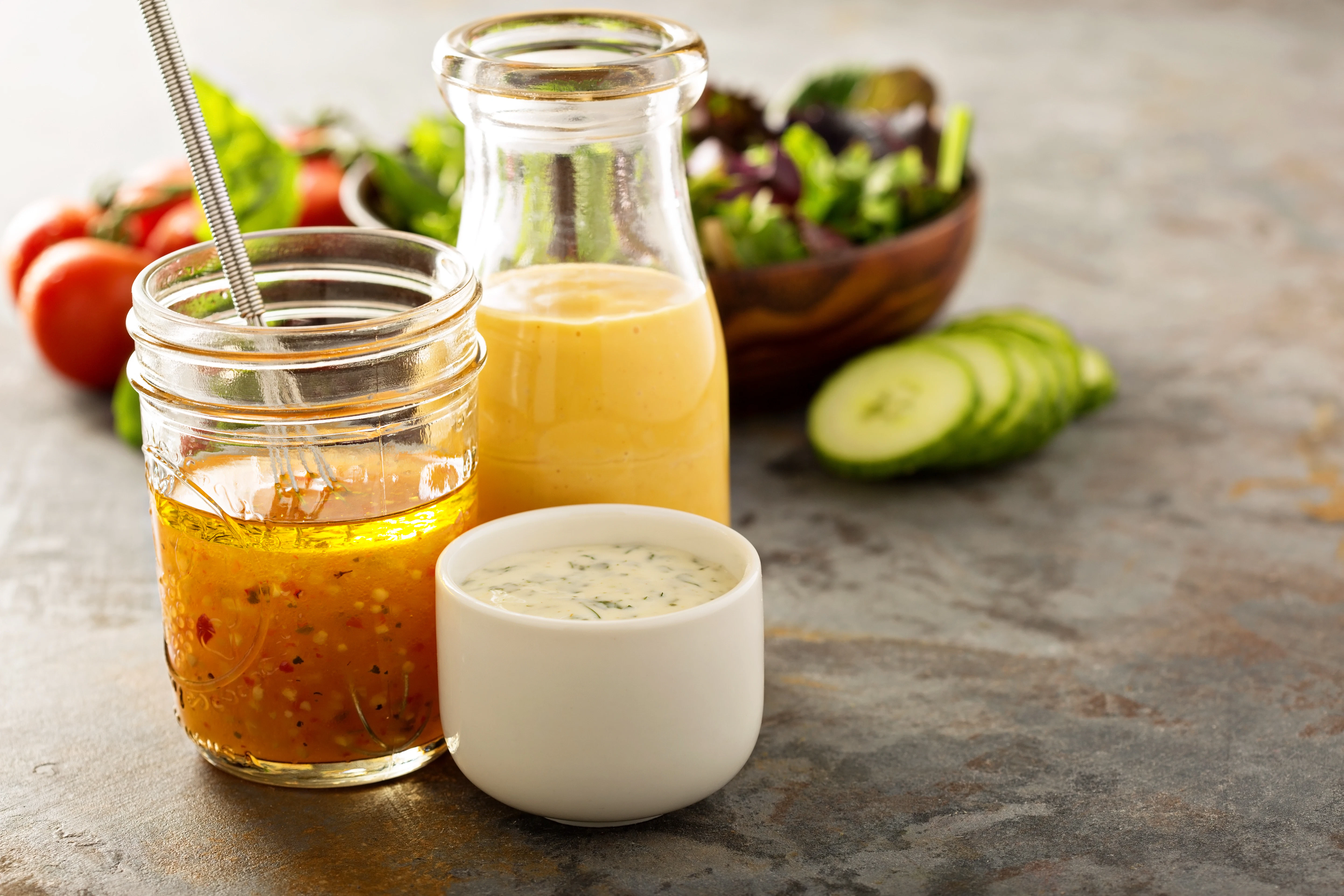 Sauces and dressings with fresh ingredients A jar of vinaigrette, a glass bottle of dressing, and a bowl of creamy sauce are placed on a table. Fresh ingredients such as cucumber slices, tomatoes, and salad leaves are visible in the background.