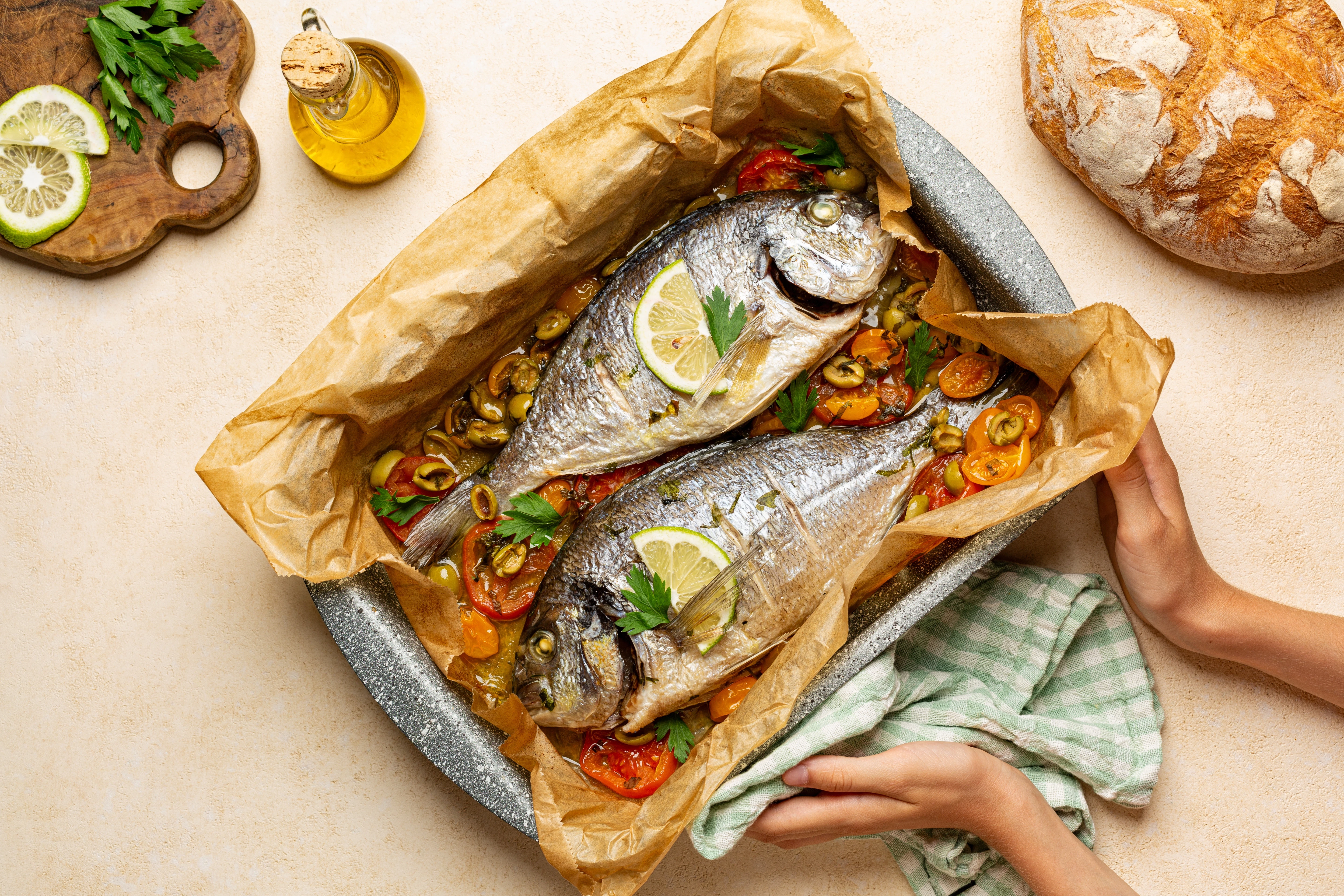 Baked whole fish with vegetables and herbs Two whole fish are baked in parchment paper with tomatoes, olives, and herbs. Lemon slices are placed on top, and hands hold the baking tray.