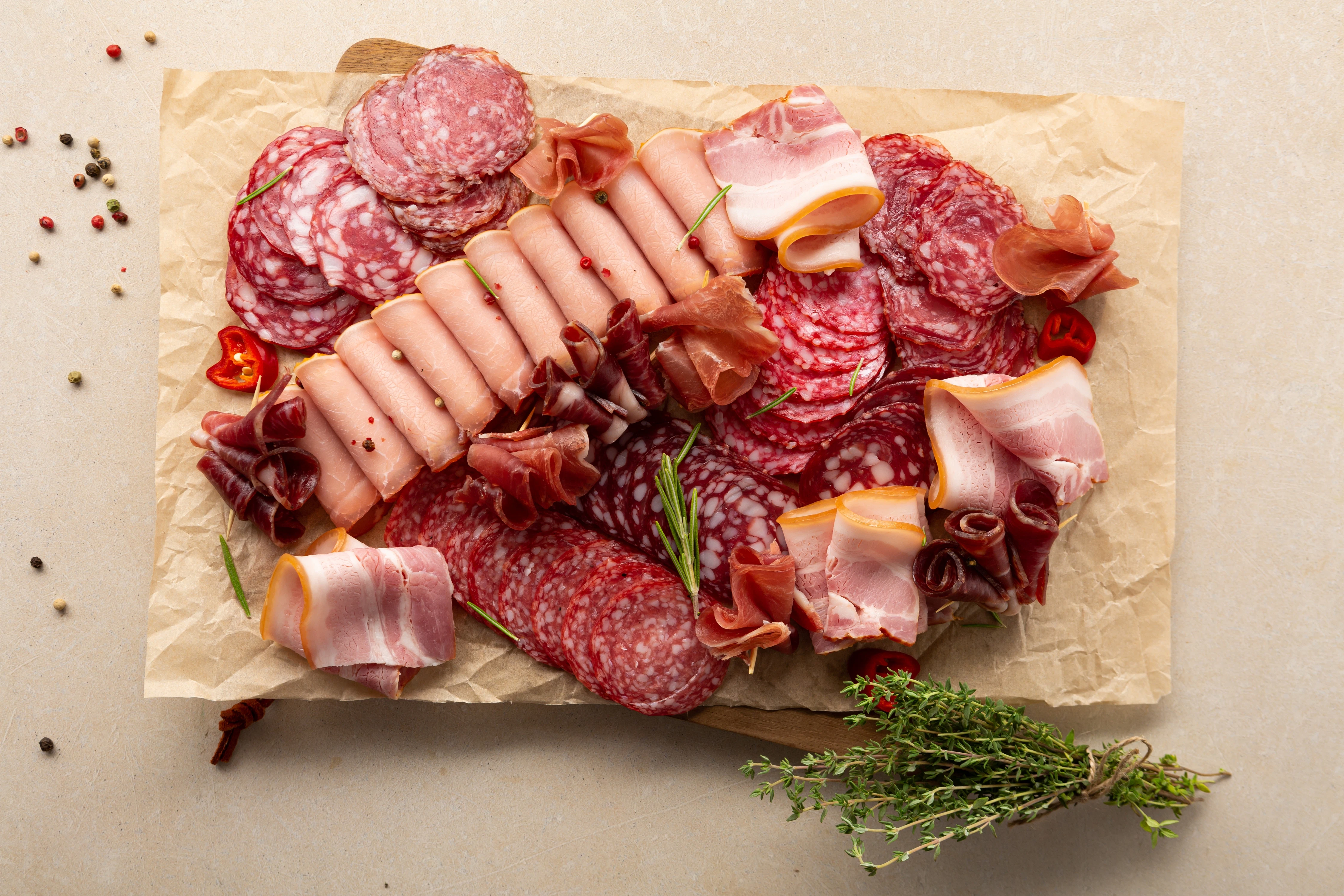 Assorted cold cuts with herbs and spices A variety of sliced meats such as salami, ham, and bacon are arranged on parchment paper. Herbs, peppercorns, and chili slices are placed around the selection.