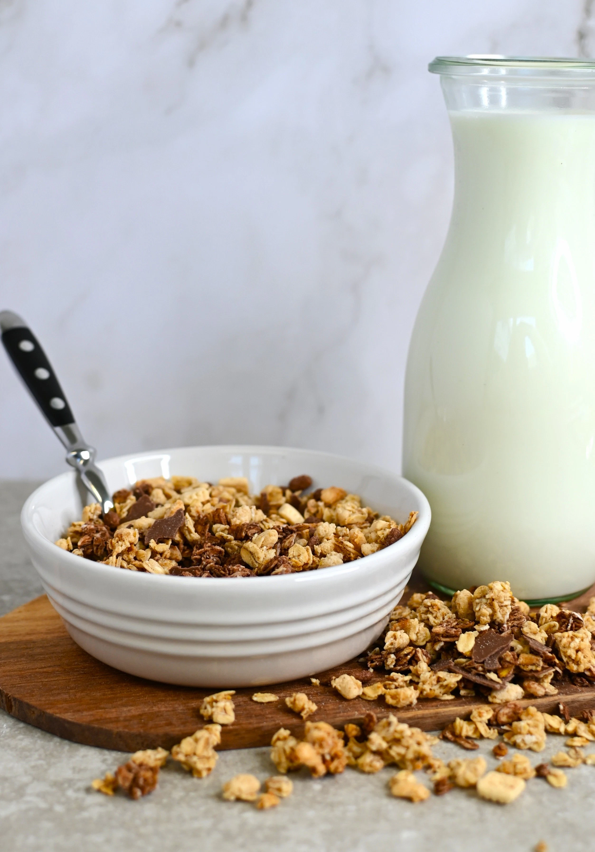 Granola with milk in a bowl and bottle A bowl of granola with nuts and chocolate pieces stands next to a glass bottle of milk. Some granola is scattered on the wooden surface.