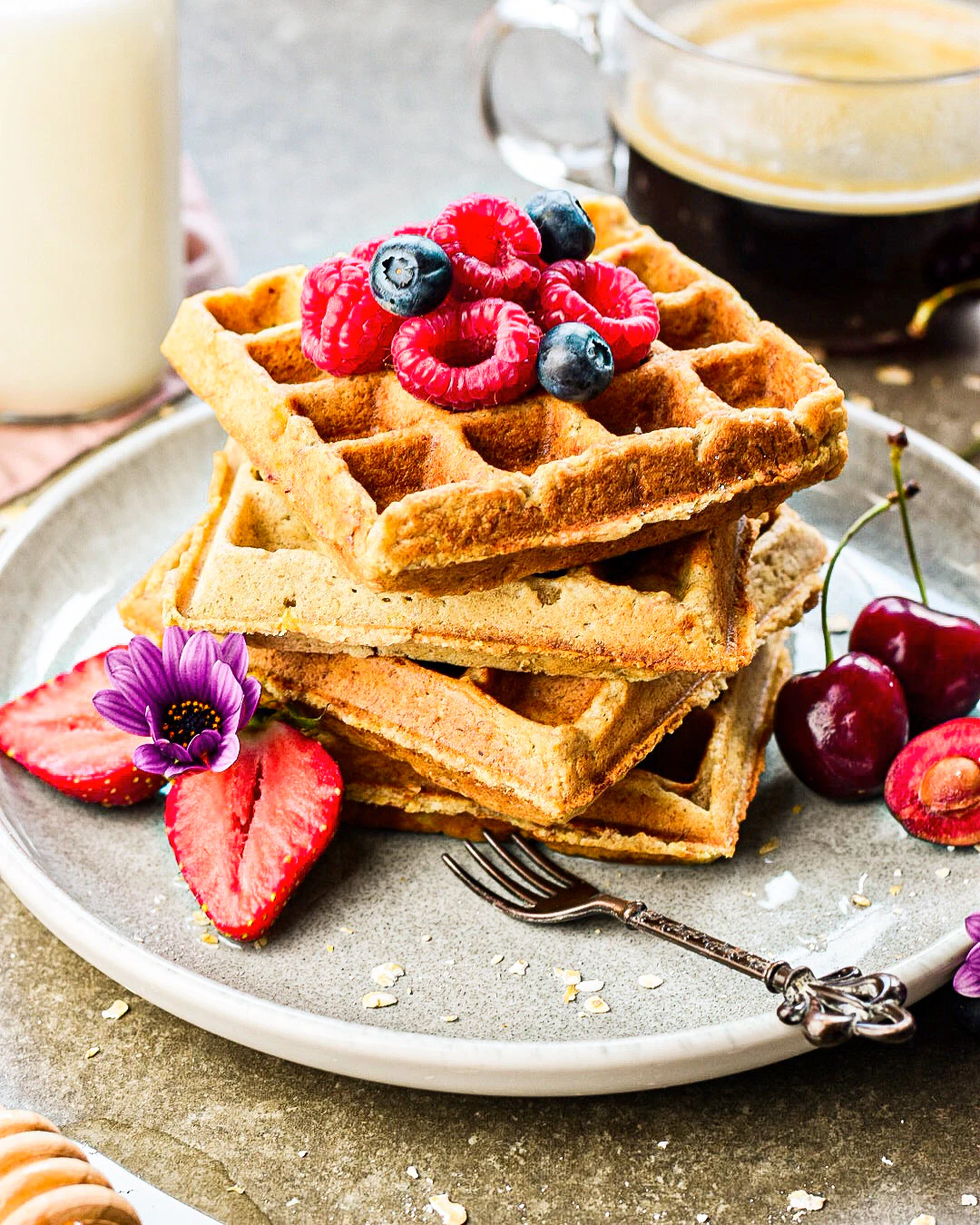 Homemade waffles with berries and cherries A stack of waffles is topped with raspberries and blueberries, surrounded by strawberries and cherries. A fork and decorative elements are placed nearby.