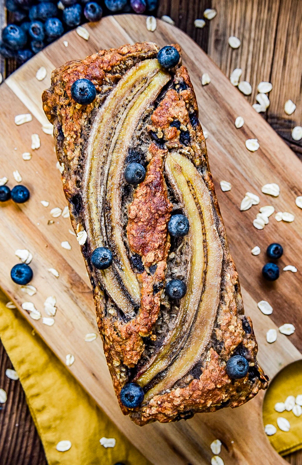 Banana bread with blueberries and oats A loaf of banana bread topped with banana slices and blueberries is placed on a wooden board. Oats and berries are scattered around the bread.