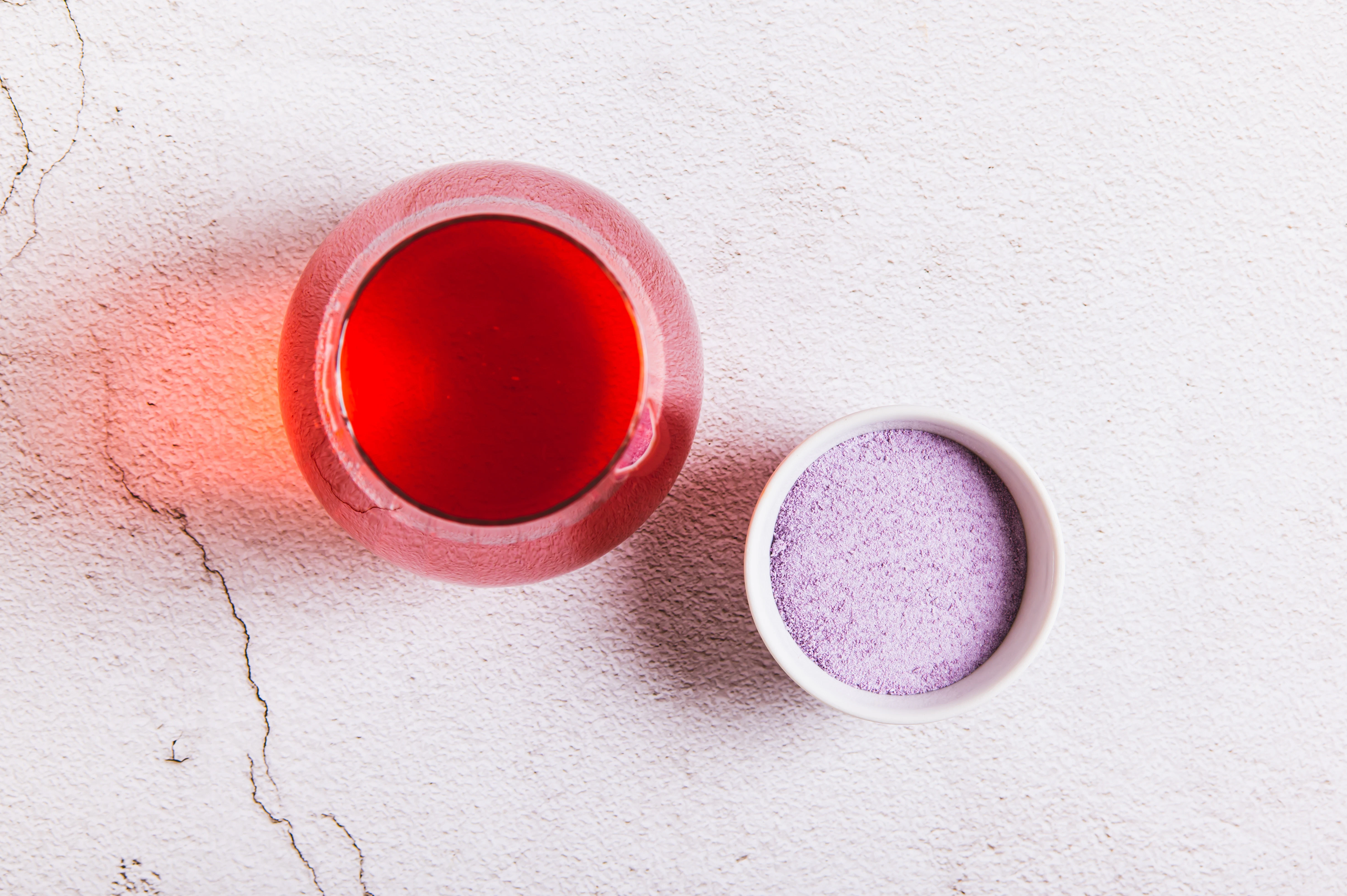 Fruit drink with powder ingredient A small glass container with red liquid stands next to a bowl of fine powder. The composition is arranged on a textured light surface.