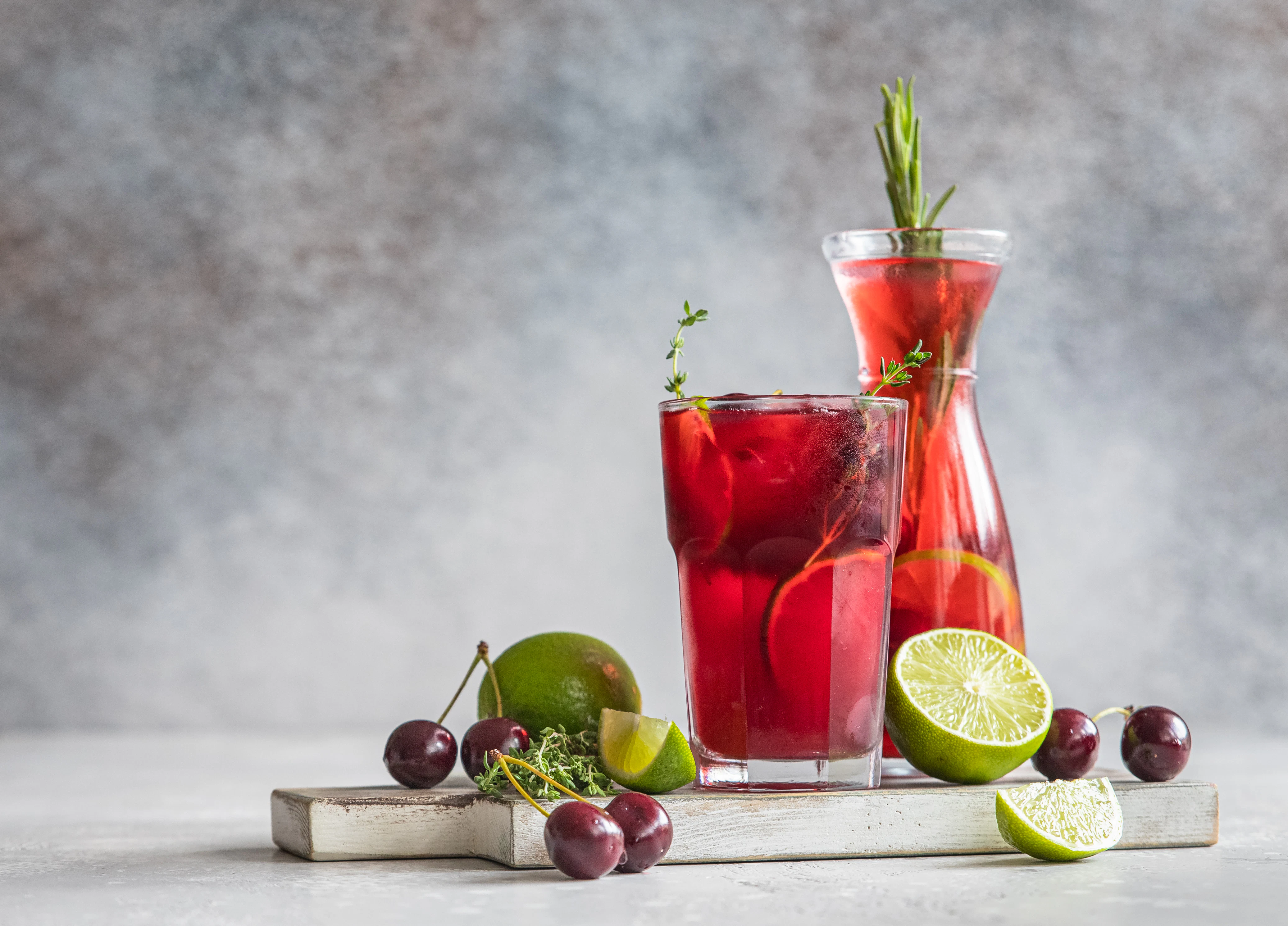 Red fruit beverage with citrus and herbs A glass and bottle filled with a red beverage are displayed alongside lime slices and cherries. Fresh herbs and ice cubes add a refreshing appearance.