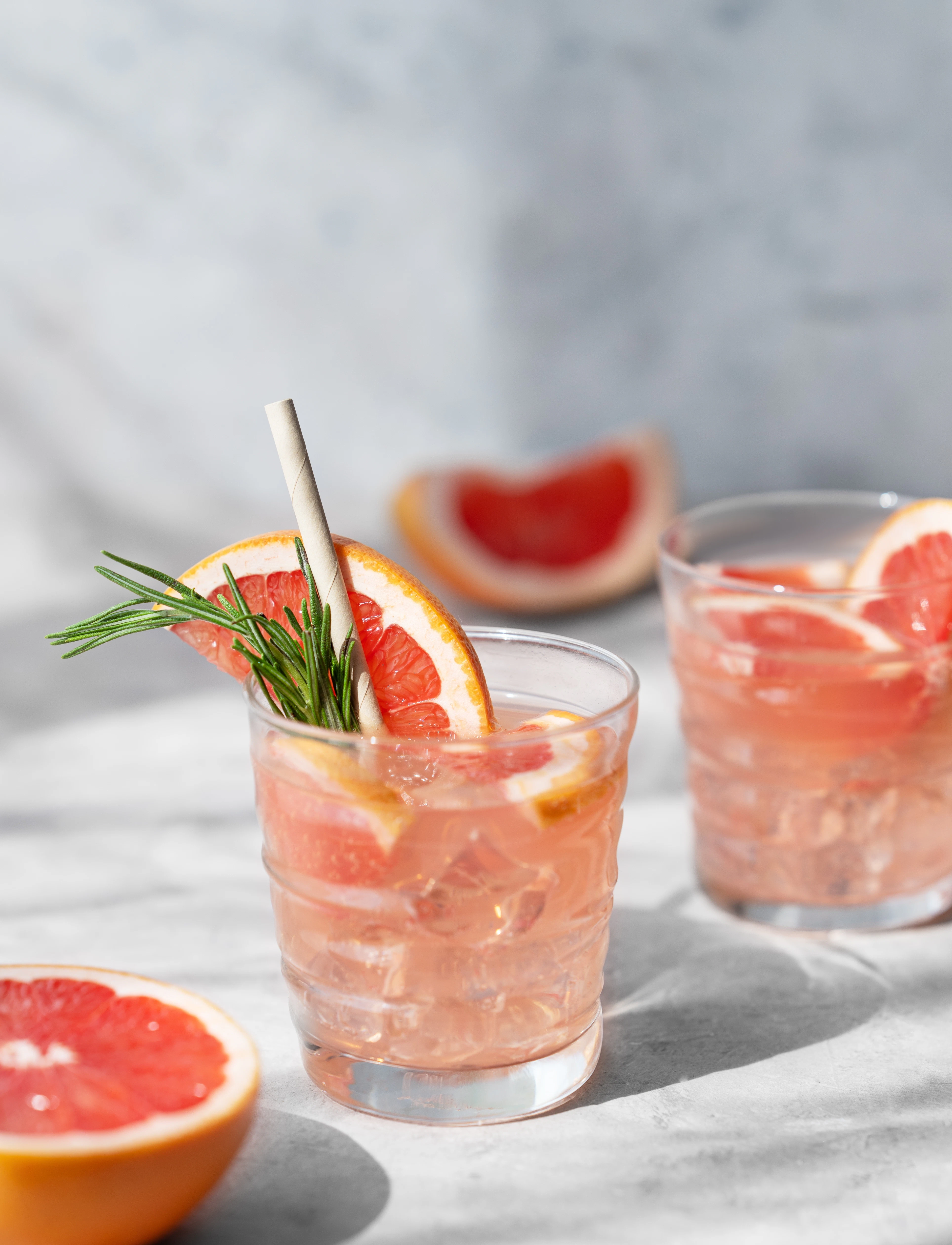 Refreshing grapefruit cocktail with rosemary Two glasses of a light pink drink are garnished with grapefruit slices and rosemary sprigs. The beverages are served on a bright surface in natural daylight.
