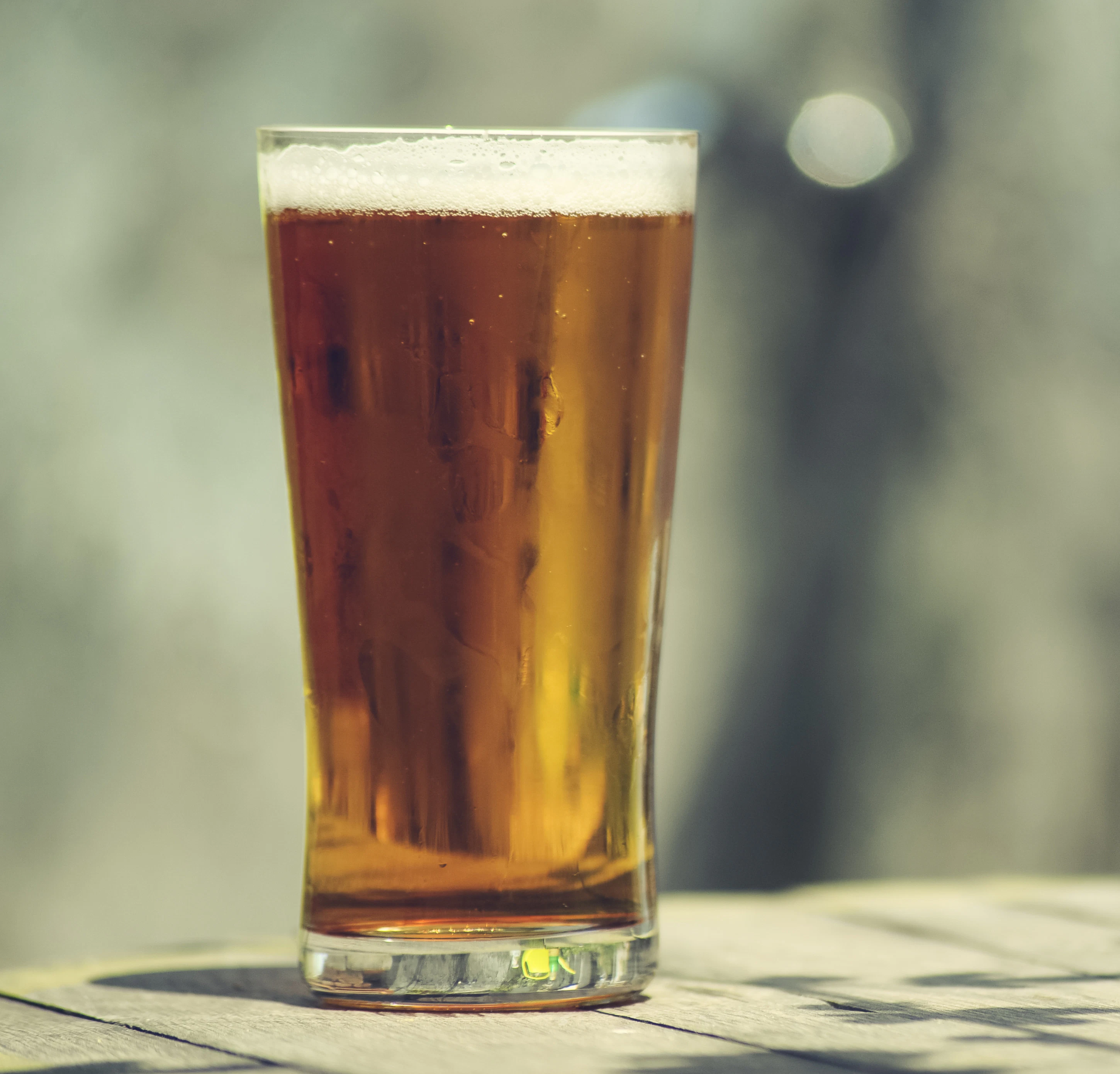 Glass of beer in sunlight A tall glass filled with beer stands on a wooden surface outdoors. Sunlight creates soft reflections around the drink.
