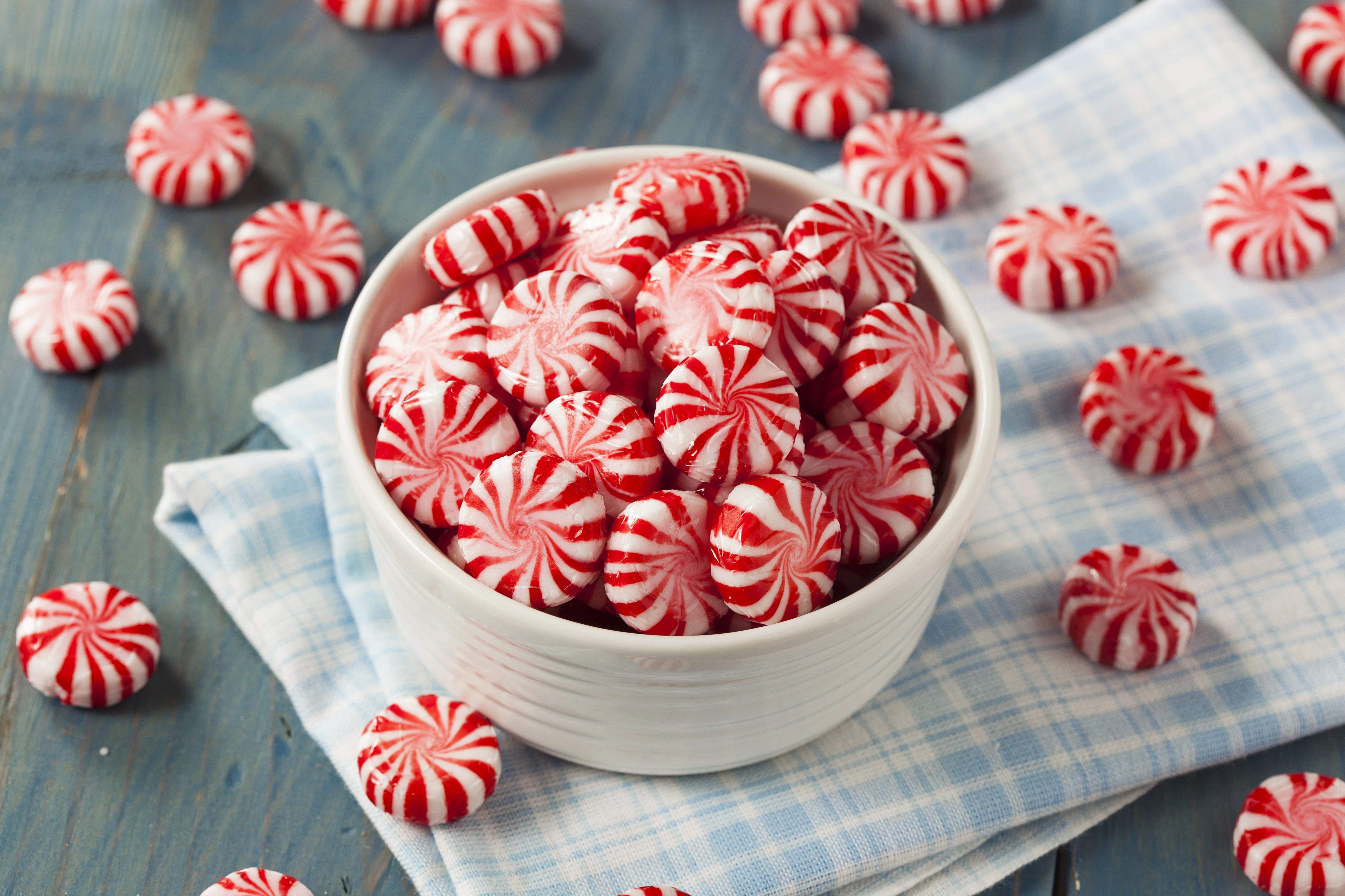 Red and white peppermint candies in a ceramic bowl A white ceramic bowl filled with red and white striped peppermint candies stands on a blue wooden surface. Several candies are scattered around the bowl on a light blue cloth and directly on the table.