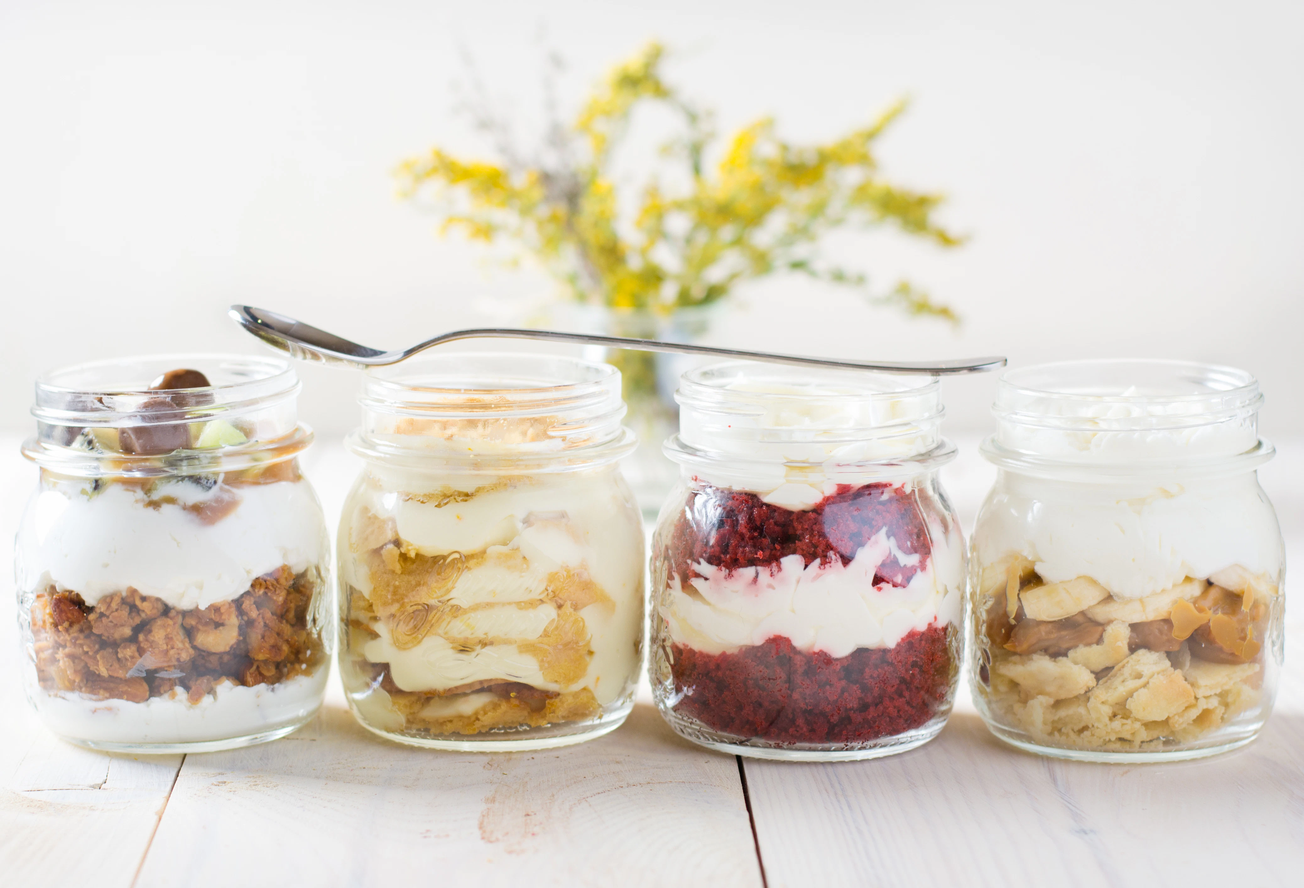 Dessert jars with layered cream fillings Four glass jars contain layered desserts with cream, cake pieces, fruit, and toppings. A spoon rests across the jars on a wooden table.