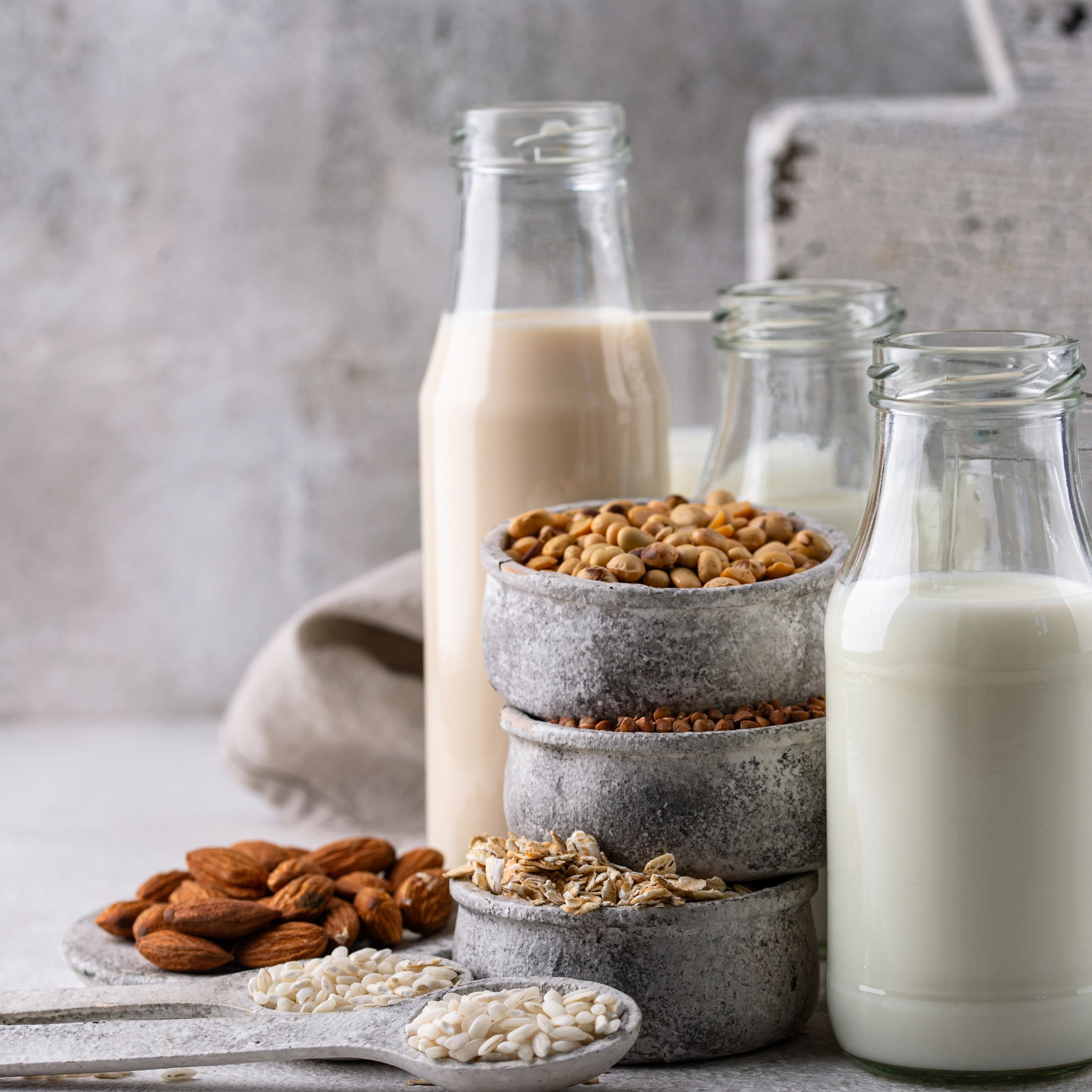 Assortment of plant-based milk alternatives with grains and nuts Several glass bottles filled with plant-based milk stand on a light surface. A stone bowl with soybeans, along with almonds, oats, and grains, are arranged in the foreground on a board and spoons, with a neutral background.