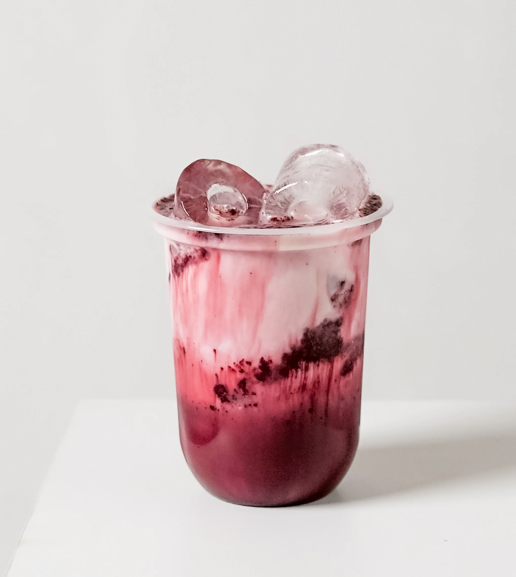Iced milk drink with berry swirl and crushed fruit A glass filled with an iced milk-based drink stands on a white surface. The beverage shows red berry swirls mixed into the milk, with crushed fruit visible and several ice cubes placed on top.