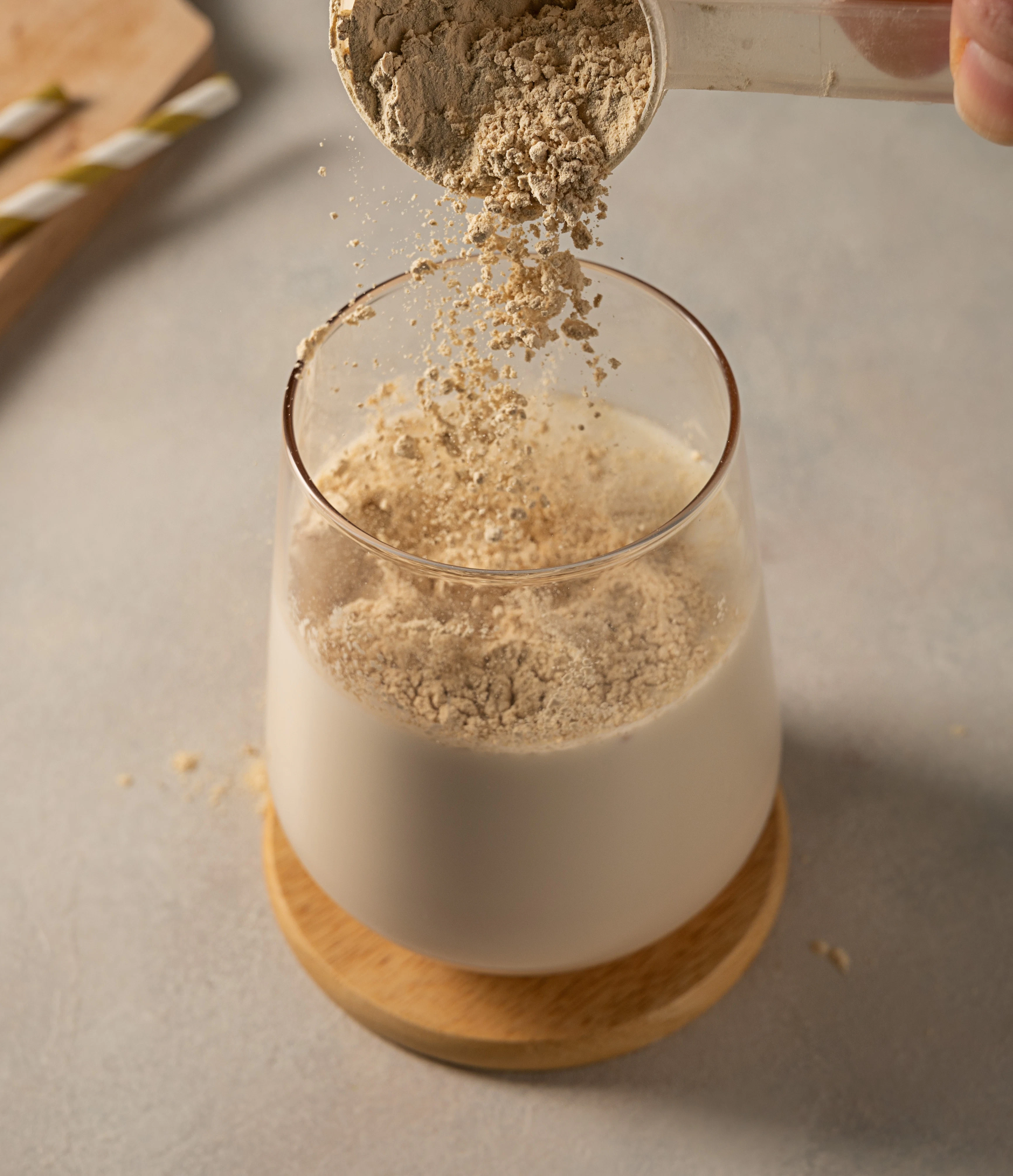 Milk-based drink with added powdered mix A glass filled with milk stands on a light surface while a scoop pours beige powder into the drink. The powder falls into the glass and begins to mix, with straws and a wooden board visible in the background.