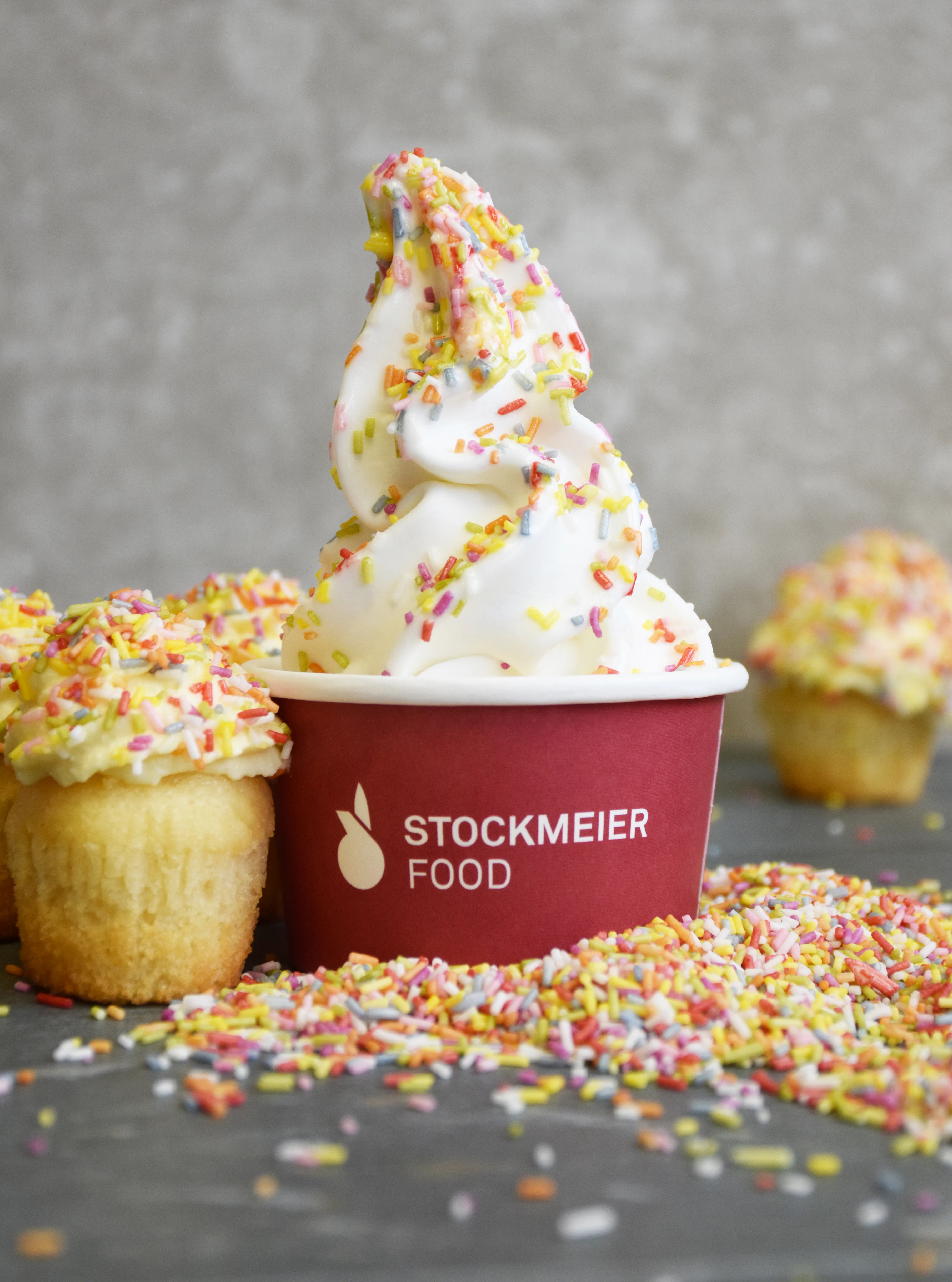 Soft serve ice cream with colorful sprinkles in a cup A swirl of soft serve ice cream topped with colorful sprinkles is served in a red cup. Cupcakes with icing and sprinkles are placed nearby, with loose sprinkles scattered on the surface.