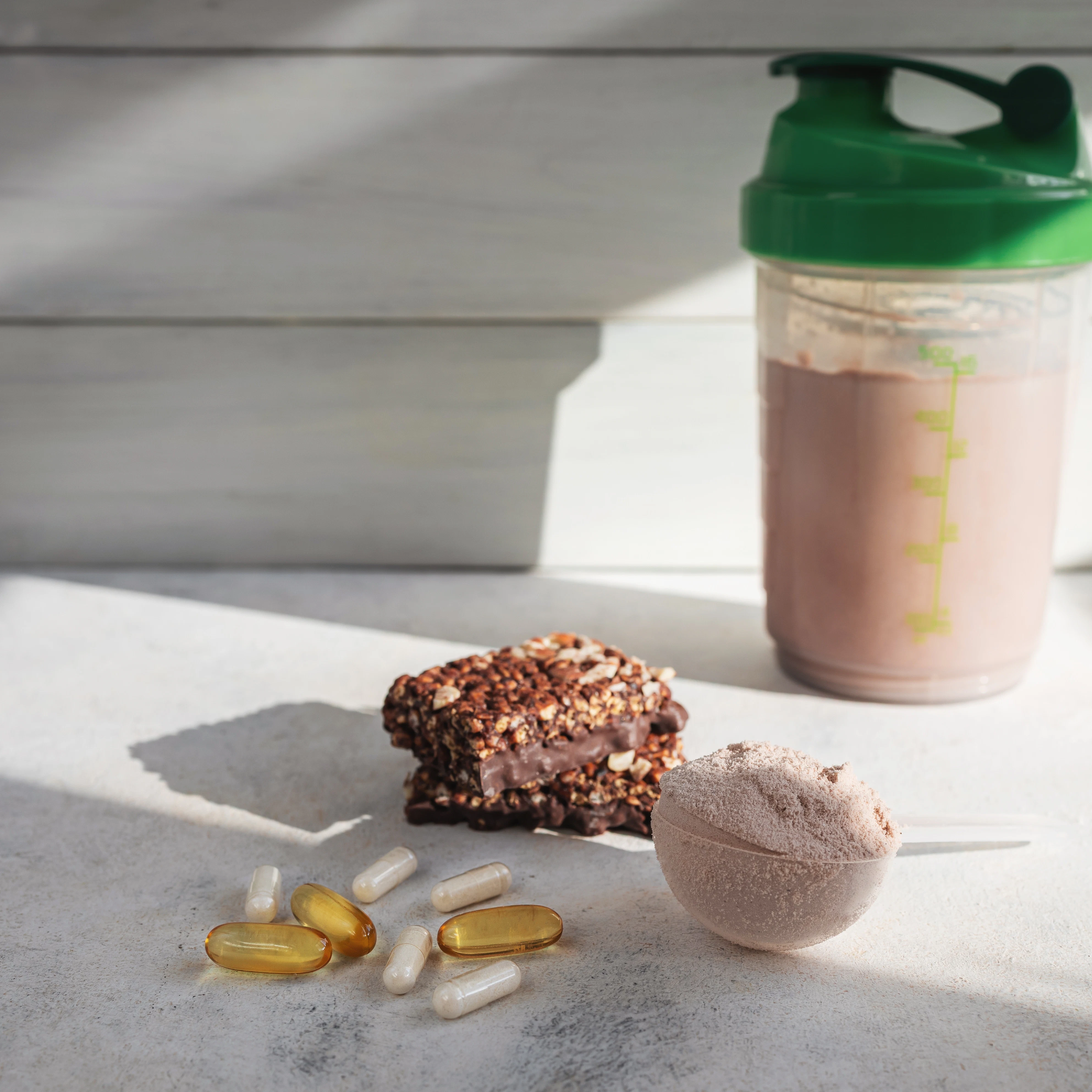 Protein shake with snack bar and supplement powder A shaker bottle filled with a light brown protein drink stands on a surface. Nearby are a snack bar, several capsules, and a scoop filled with powder.