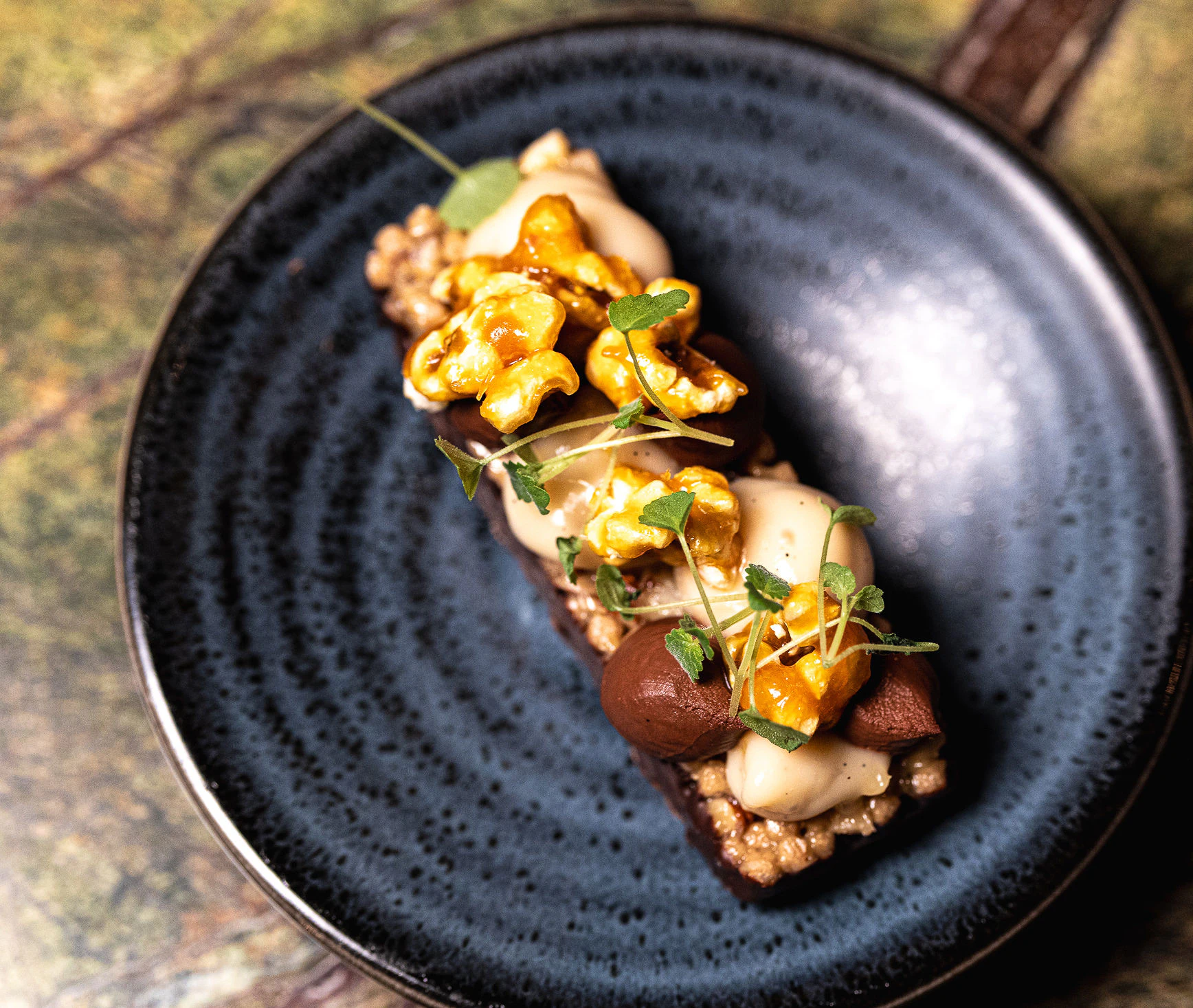 Premium dessert creation with caramelized nuts A plated dessert is decorated with caramelized nuts, cream elements, and microgreens. The dish is presented on a dark ceramic plate.