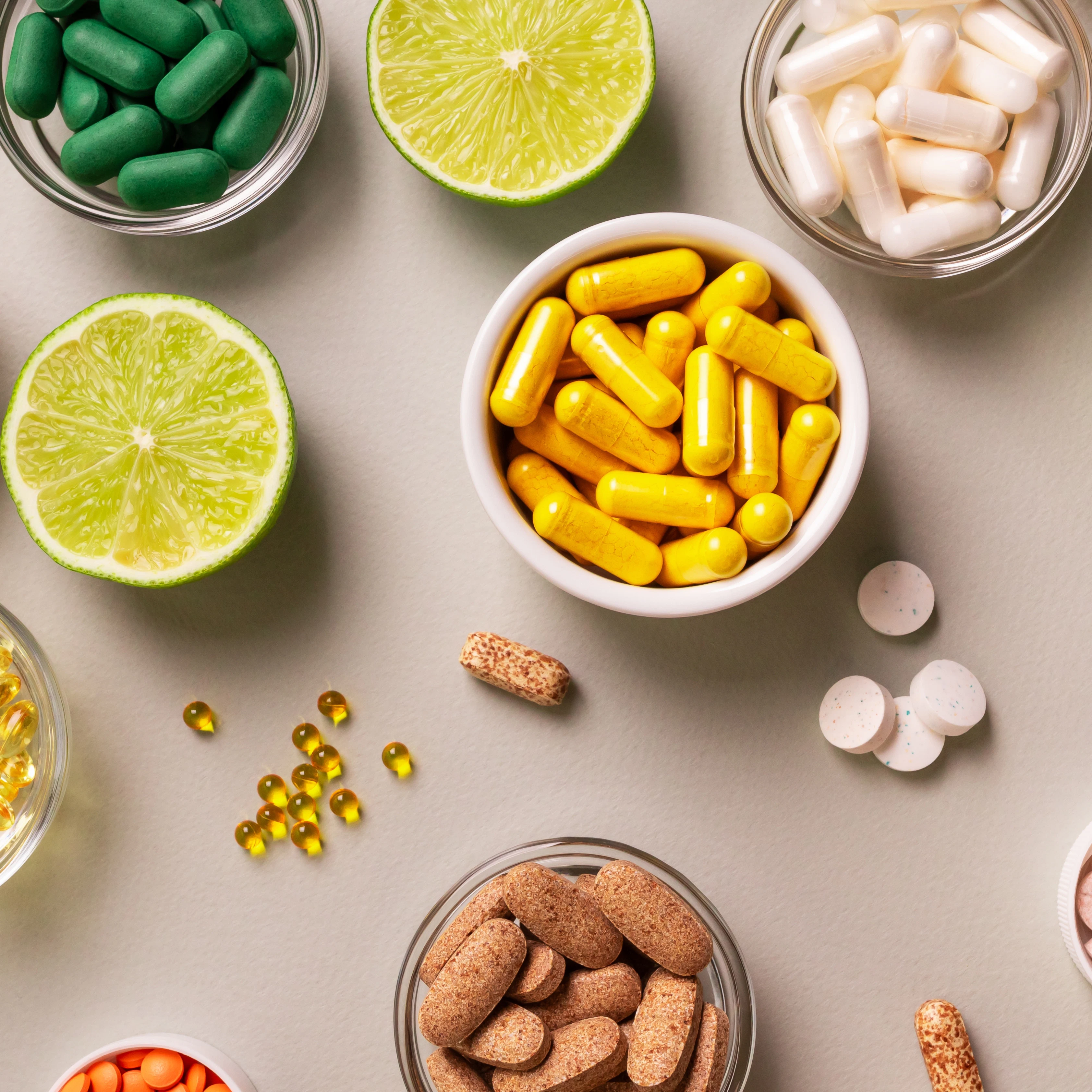 Assorted dietary supplements with citrus fruits Bowls filled with capsules and tablets in different colors are arranged on a light surface alongside halved limes. Additional pills and soft gel capsules are placed around the bowls.