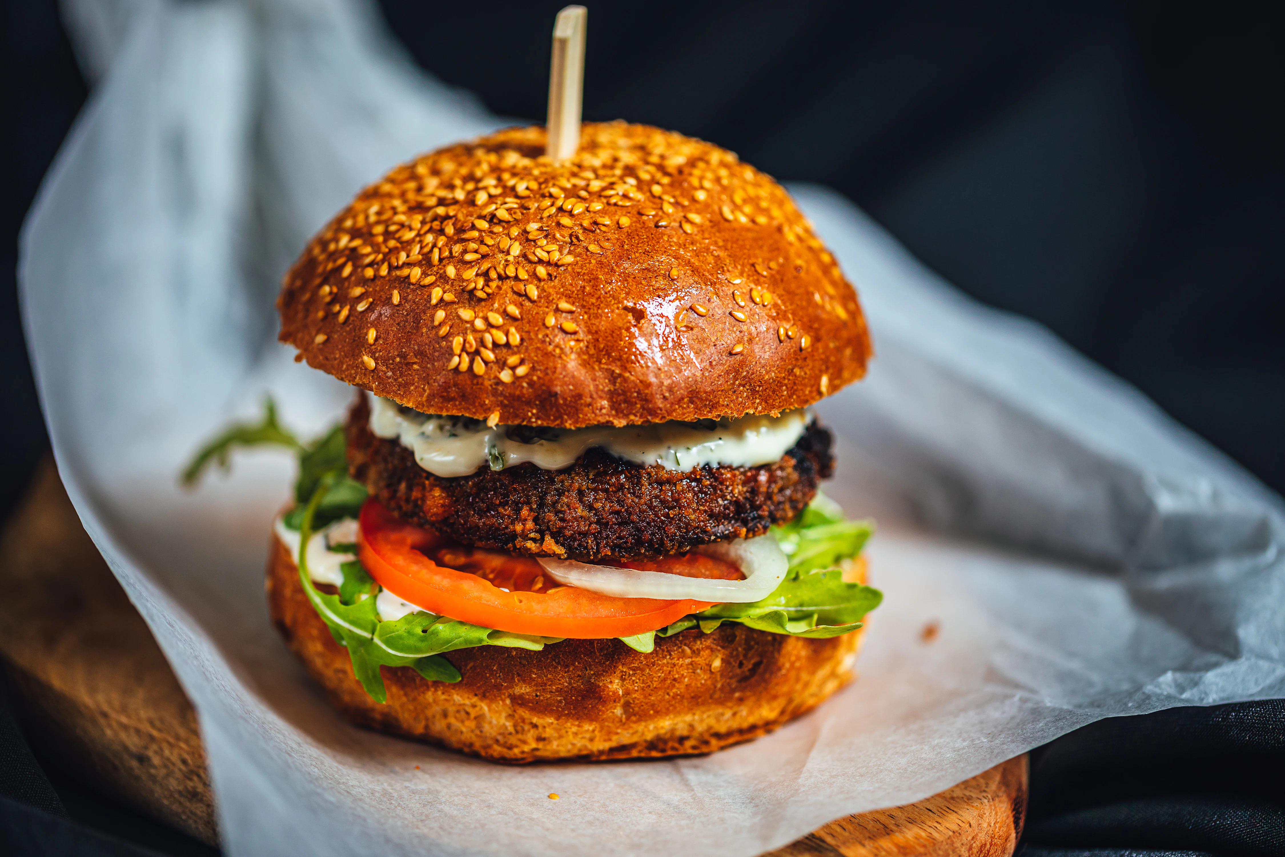 Gourmet burger with crispy patty and fresh toppings Close-up of a burger with a sesame bun, crispy fried patty, lettuce, tomato, onion, and creamy sauce. The burger is placed on parchment paper on a wooden surface.