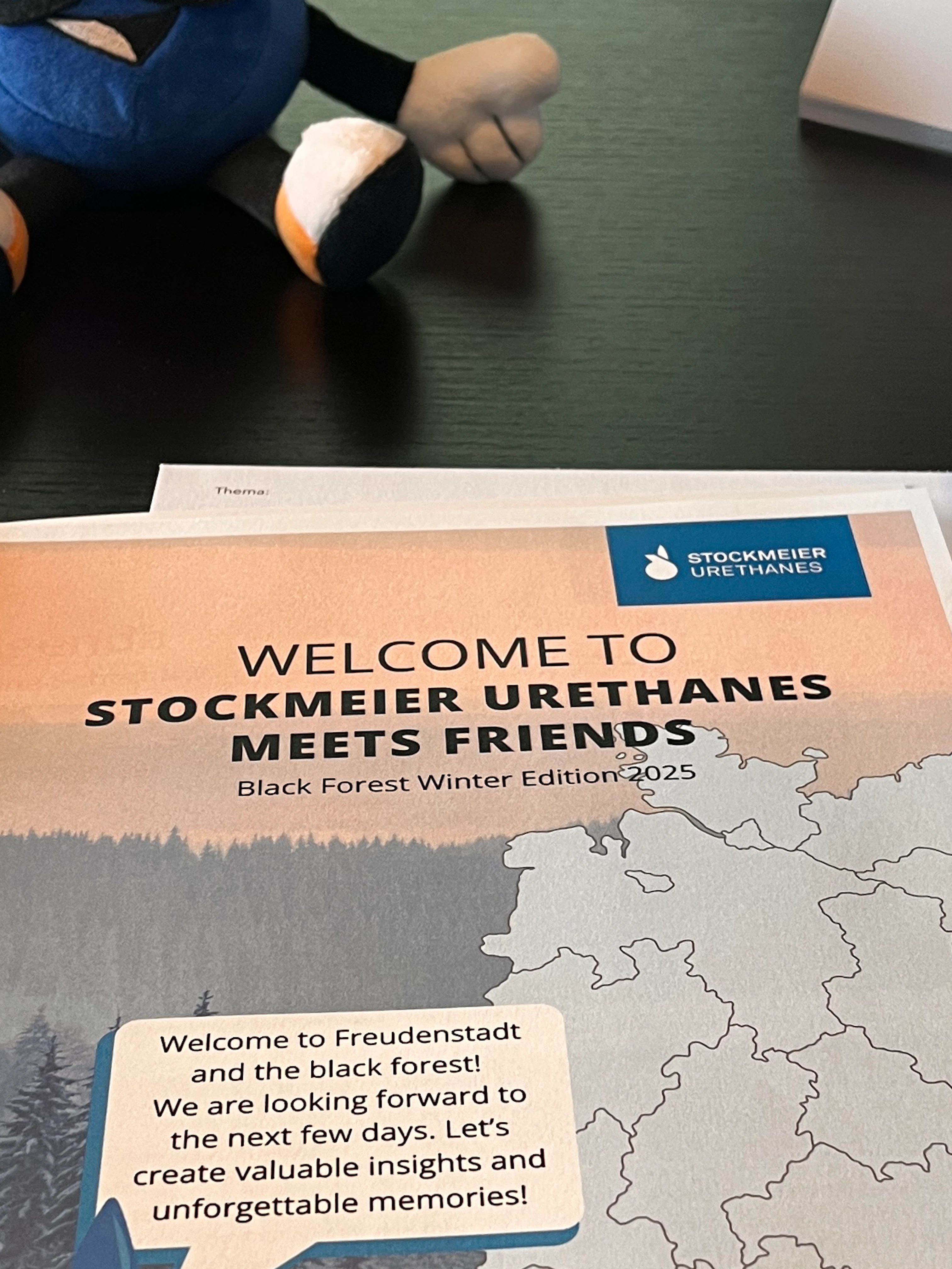 Stobi neben der Willkommensmappe Stobi sizt hinter einem Blatt, dass alle Teilnehmenden beim STOCKMEIER Urethanes Meets Friends event willkommen heißt