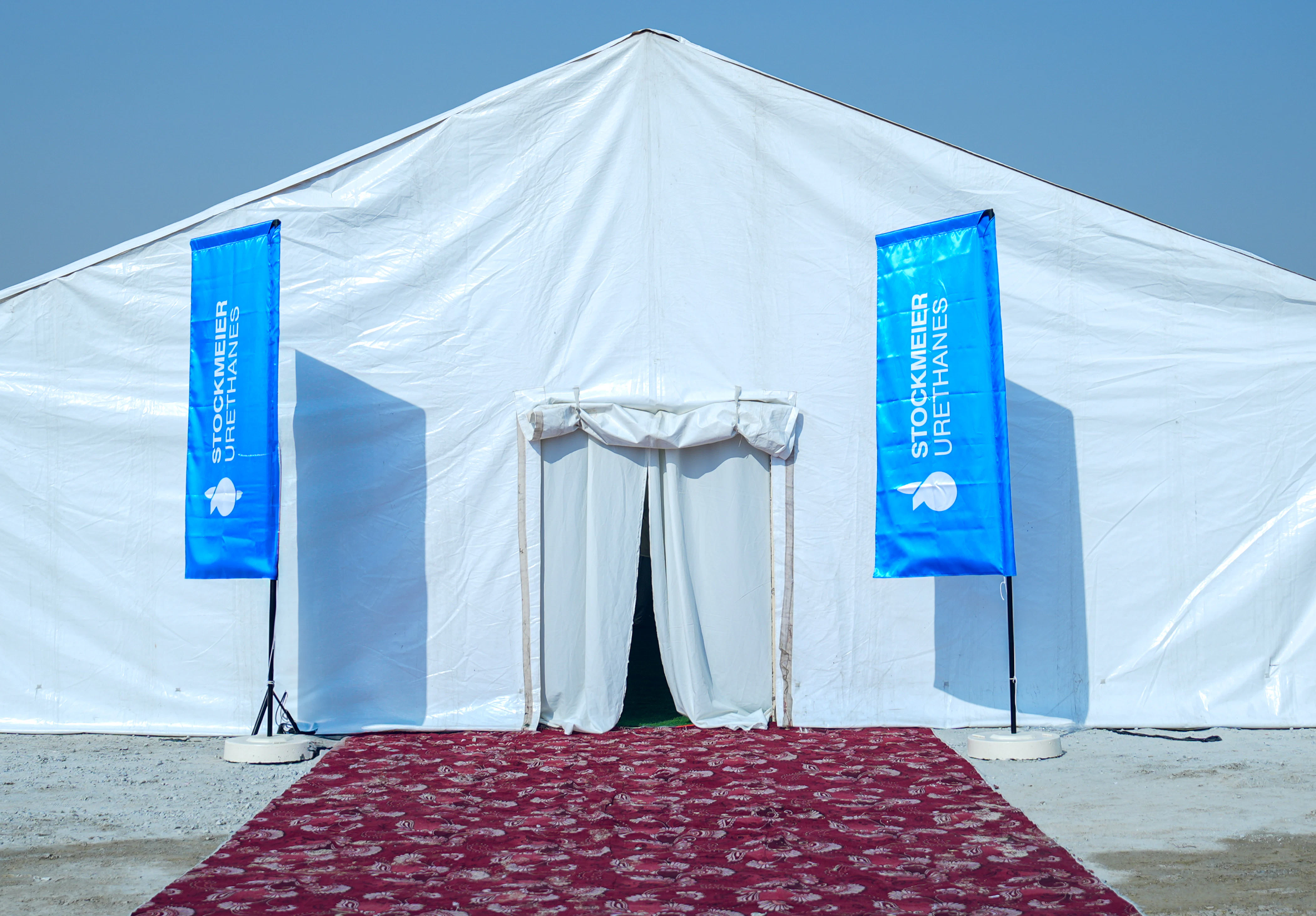Zelt der Spatenstich Zeremonie von STOCKMEIER Urethanes in Dubai Vorderansicht des Zelts mit einem roten Teppich und zwei blauen Strandlaggen mit Firmenlogo neben dem Eingang